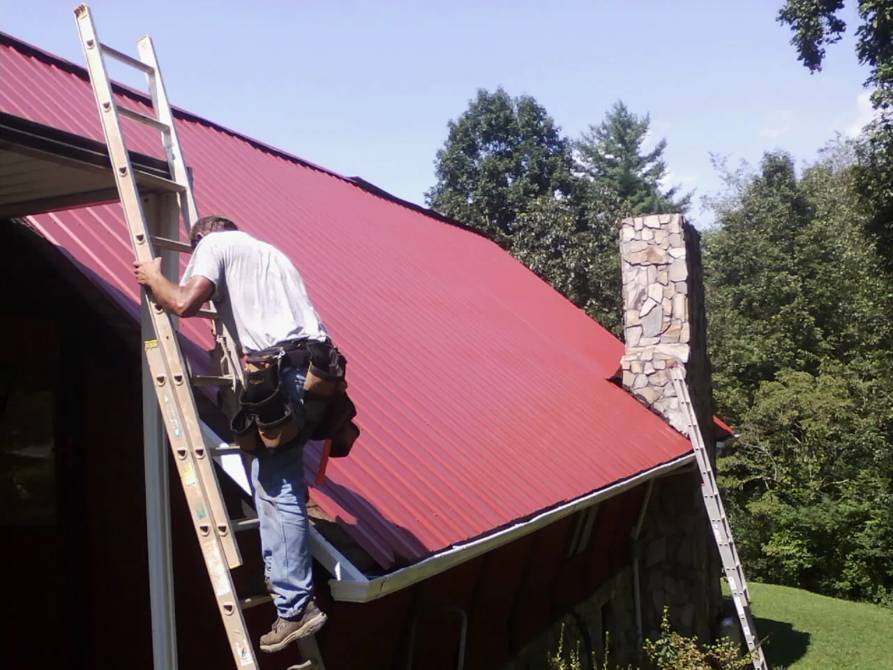 Roofer on a ladder beside a red metal roof and stone chimney — Soffit & Fascia Repair by Roofing Brothers in Salt Lake City